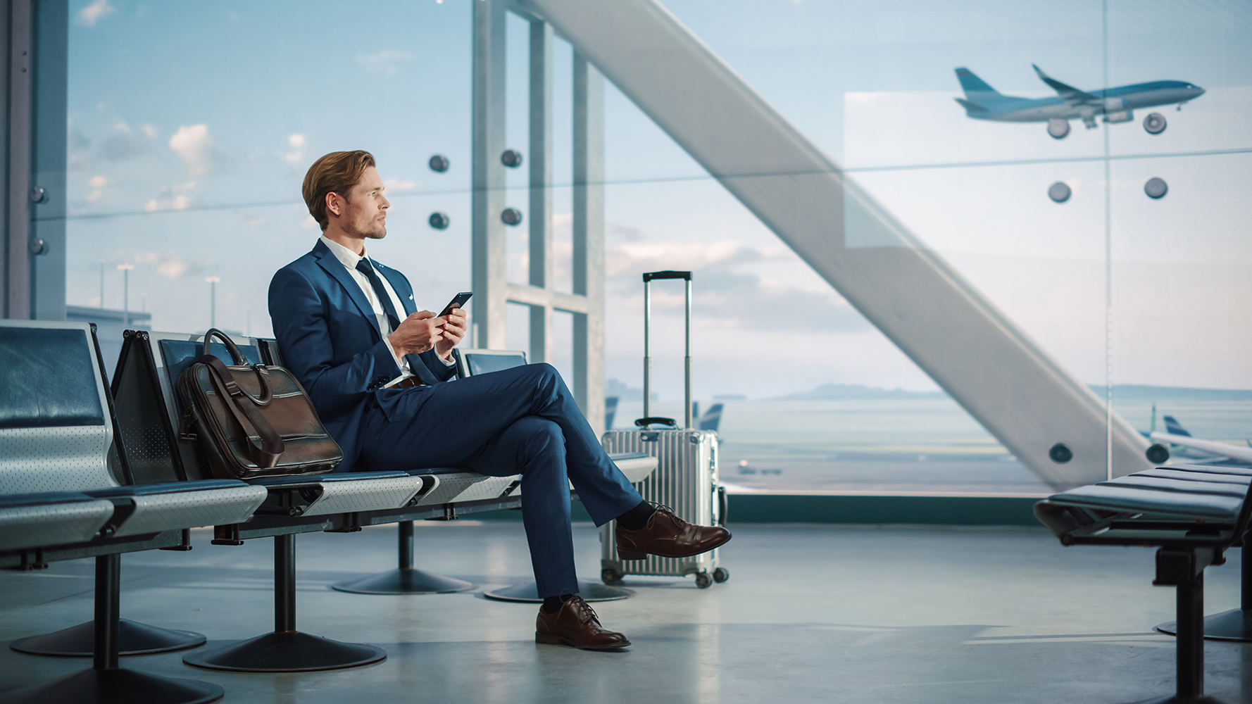 Businessman in a blue suit sitting in an airport lounge, using a smartphone, with a suitcase nearby and an airplane visible through a large window, reflecting a comfortable and professional travel experience.