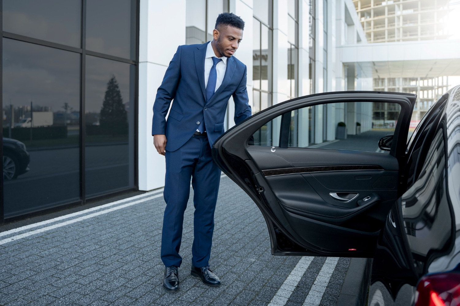 Professional chauffeur in a suit opening the door of a luxury sedan outside a modern building, emphasizing premium transportation services in Spring, Texas.